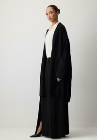 Garšs melns kardigans, izgatavots no mīksta auduma, ar atvērtu priekšpusi, platām piedurknēm un brīvu piegriezumu, uzvilkts pāri gaismas krāsas blūzei un garajām svārkiem.