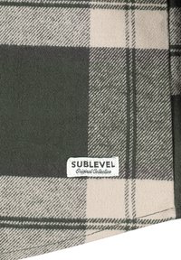 Grün-beige Karostoff mit weicher Textur, an der Kante mit einem Etikett "SUBLEVEL Original Collection" genäht.