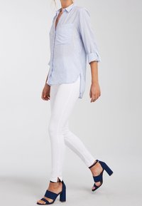 Femme marchant, portant une chemise à rayures bleu clair à manches longues, un pantalon blanc ajusté et des sandales à talons hauts bleu marine avec des brides de cheville.