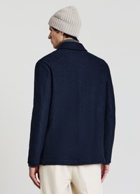 Giacca in pile blu navy con colletto e vestibilità dritta, caratterizzata da una superficie testurizzata e tasche laterali. Indossata con un berretto a coste beige chiaro.
