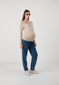 Beige ribbed off-shoulder knit sweater, pregnant model wearing loose blue denim jeans and beige sneakers, with rectangular sunglasses.