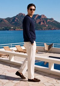 Homme portant des lunettes de soleil, un pull bleu marine, un pantalon blanc et des chaussures marron marchant sur une terrasse au bord de la mer avec des montagnes et des chaises longues en arrière-plan.
