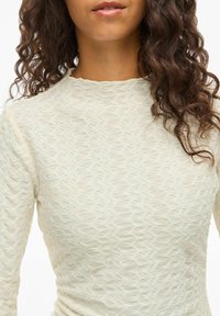 Helles beige Oberteil aus strukturiertem Stoff mit welligem Muster, Rundhalsausschnitt und langen Ärmeln, das eine taillierte Silhouette betont.