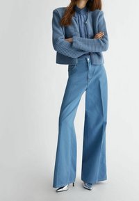 Vaaleansininen, teksturoitu neulejakku, jossa on lyhyt malli ja napit, yhdistetty korkeavyötäröisiin, levenevin lahkein vaaleansinisiin farkkuihin ja hopeisiin, kärjestä teräviin korkokenkiin.