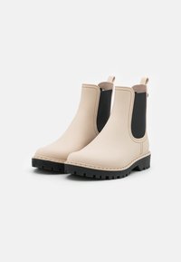 Botas de tobillo de goma beige con paneles laterales elásticos, suelas de goma negras y costuras reforzadas. Presenta tiradores para facilitar su uso.