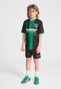 Svart och grönrandig sporttröja med logotyp och text, tillsammans med svarta shorts, vita strumpor och gröna sneakers med kardborreband.