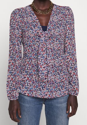 Person med mørk hud, iført en navyblomstret bluse med lange ærmer, bind-foran detalje, blå undertrøje, guldkæde halskæde og blå jeans.