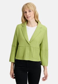 Zielona, strukturalna marynarka z wykładanym kołnierzem, dwoma przednimi kieszeniami i rękawami o długości ¾, w połączeniu z białą bluzką i czarnymi dżinsami.