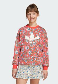 Červená kvetinová mikina s bielym logom Adidas, okrúhly výstrih a rebrované manžety, v kombinácii s bielou sukňou s kvetinovým vzorom.