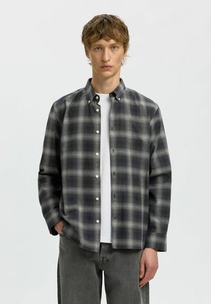Jeune homme aux cheveux châtain clair courts porte une chemise à carreaux noire et grise à boutons par-dessus un t-shirt blanc et un pantalon gris foncé, debout de manière neutre.