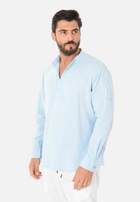 Camicia di lino blu chiaro con colletto alla cinese, caratterizzata da un'apertura con bottoni e maniche lunghe con polsini abbottonati. Tessuto morbido.