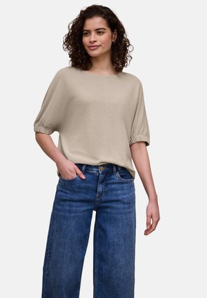 Donna con capelli ricci che indossa una maglietta beige a maniche corte larga e jeans blu a gamba larga a vita alta, in piedi con una mano in tasca.