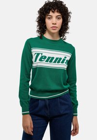 Pull en tricot vert avec le texte "Tennis" en blanc, présentant des poignets et un ourlet côtelés avec des rayures blanches. Associé à un pantalon en denim foncé.