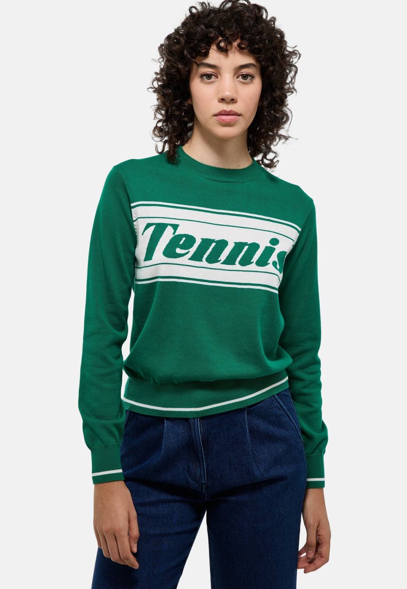 Pull en tricot vert avec le texte "Tennis" en blanc, présentant des poignets et un ourlet côtelés avec des rayures blanches. Associé à un pantalon en denim foncé.