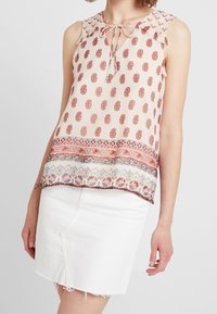 Blusa senza maniche in pesca chiaro con motivo paisley marrone, dettagli in pizzo all'orlo e scollo a legatura. Abbinata a una gonna di jeans bianca.