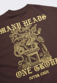 T-shirt di cotone marrone con stampa grafica in oro di un drago a molte teste sopra "MOLTE TESTE" e "UN SOLO GRUPPO", con dettagliati lavori a linee.