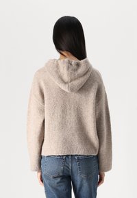 Felpa a collo alto in maglia beige realizzata con un tessuto morbido e texturizzato; vestibilità ampia e polsini a coste; presenta un cappuccio con cordino e un design leggermente cropped.