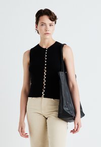 Zwart mouwloos topje met een ronde halslijn, voorzien van een rij witte knopen aan de voorkant; gecombineerd met beige broek en een zwarte totebag.