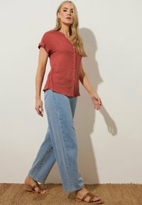 Camisa corta de manga con botones en color óxido, con escote en V, combinada con jeans de pierna ancha en azul claro y sandalias planas marrones. Fondo de alfombra de yute.