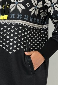 Sort hættetrøje med hvide og blå snefnug- og hjerte mønstre, med en frontlomme og kontrasterende gule snore. Blød stoftekstur.