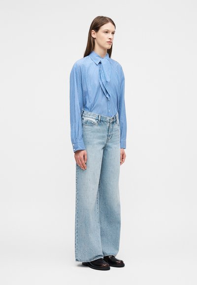 Γυναίκα στέκεται φορώντας μπλούζα με μπλε ρίγες και βολάν στο μπροστινό μέρος, φαρδιά ανοιχτό μπλε τζιν παντελόνια με μικρές διακοσμήσεις και μαύρα παπούτσια σε λευκό φόντο.