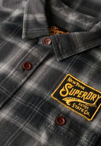 Szara i czarna flanelowa koszula w kratę z brązowymi guzikami oraz haftowaną żółtą łatką z logo Superdry na klatce piersiowej.