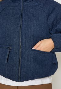 Tumši zils džinsu jakas ar rāvējslēdzēju, ribotām piedurknēm un divām sānu kabatām. Materiāls izskatās teksturēts un izturīgs.