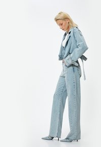 Chaqueta de mezclilla azul clara con bordes deshilachados y cierre de lazo, combinada con pantalones vaqueros de pierna ancha. Unos tacones de punta completa el look.