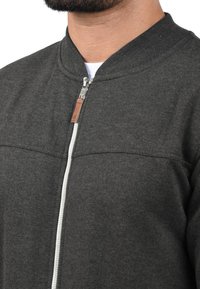 Grå zip-jacket med rund krave, der har en kontrasterende hvid lynlås og en lille læderdetalje nær lynlåshåndtaget.