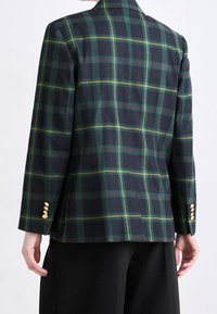 Veste à carreaux foncés avec des lignes vertes, jaunes et rouges. Tissu lisse avec un design à simple boutonnage et des boutons dorés en accent sur les manches.