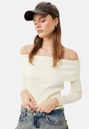 Nuori nainen, jolla on ruskea lippalakki, olkaimeton kermaisen värinen ribbineule ja siniset farkut, katsoo alaspäin kädet lähellä farkkujen vyötäröä.