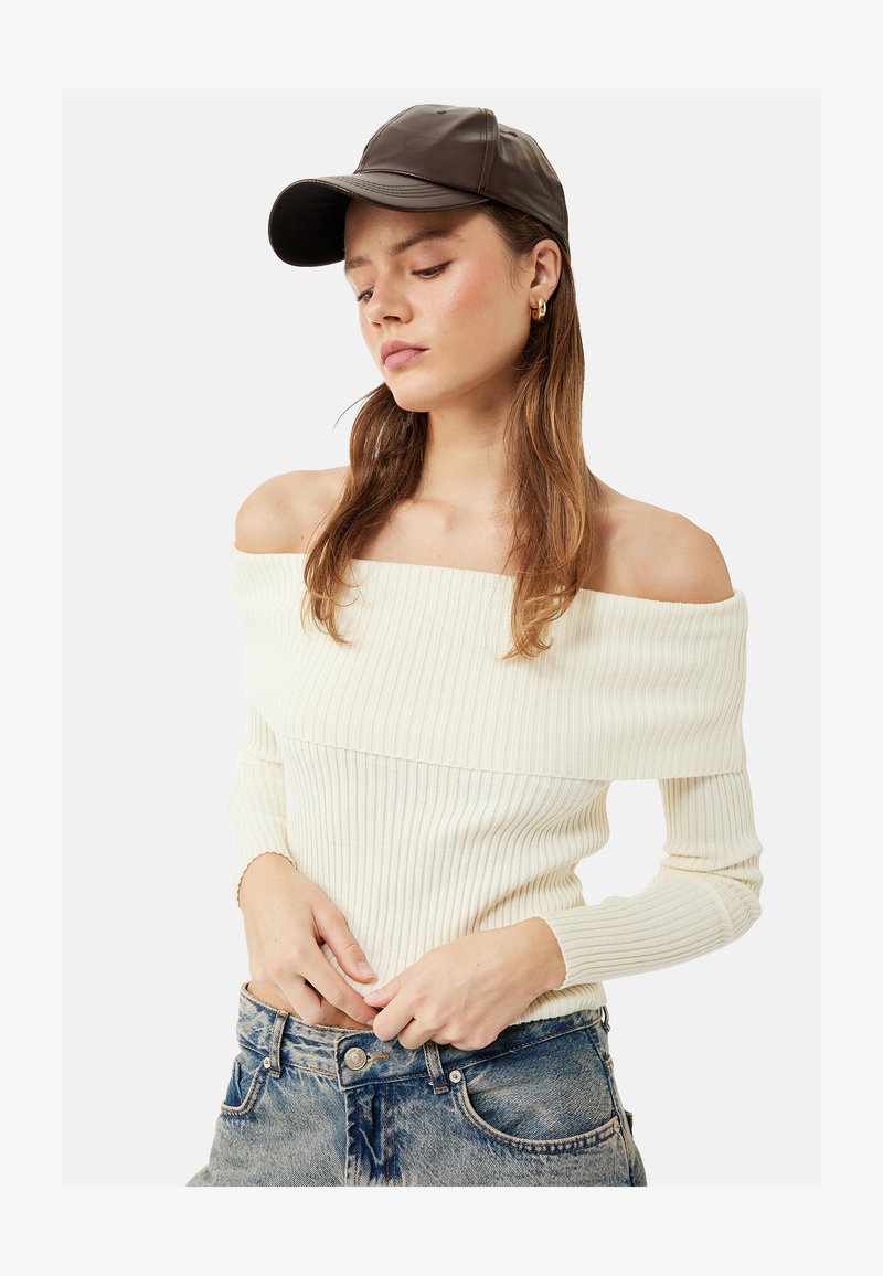 Joven mujer con gorra marrón, suéter crema de canalé sin hombros y vaqueros azules, mirando hacia abajo con las manos cerca de la cintura de los vaqueros.