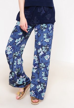 Kvinde iført løse blå bukser med stort blomsterprint i nuancer af blågrøn, marineblå og hvid, kombineret med gyldne kilehælssandaler og en mørk top.