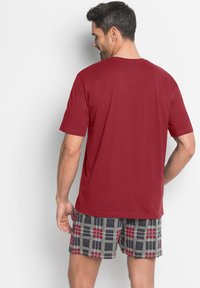 T-shirt rossa in cotone con maniche corte, vista da dietro, abbinata a pantaloni corti a quadri grigi con accenti rossi e neri.