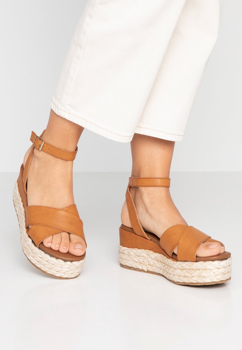 Sandales compensées en cuir marron avec des brides croisées et une boucle à la cheville, avec une plateforme texturée style jute et un design à bout ouvert.
