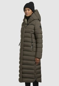 Long puffer coat in brown, features a hood, vertical quilting, side zip pockets, and snap buttons. Worn with a black knit hat and black attire.