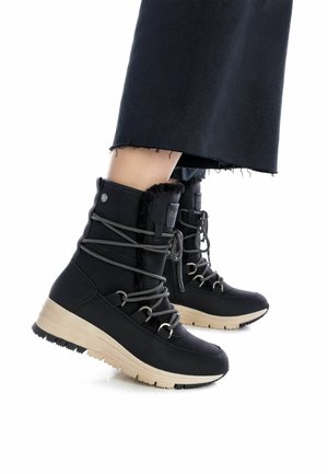 Botas aislantes negras con un acabado texturizado, presentando un caña alta, diseño de cordones y suela de goma beige. Los acentos incluyen herrajes plateados y piel sintética.