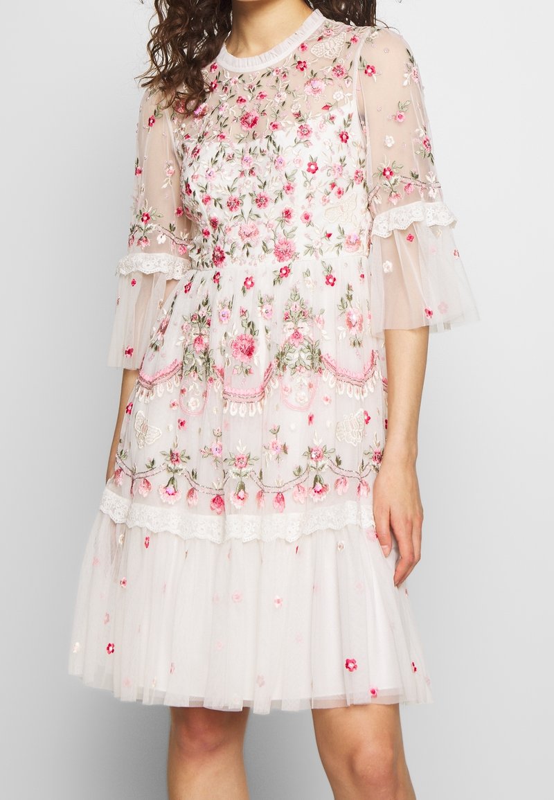 Femme portant une robe blanche mi-longue avec une broderie florale rose et des manches transparentes à volants.