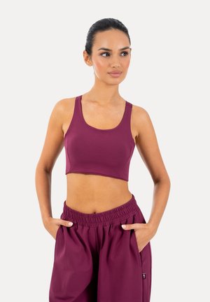 Kobieta z ciemnymi włosami związanymi w kok, ubrana w fioletowy bezrękawnik i pasujące luźne spodnie, z rękami w kieszeniach na jednolitym tle.