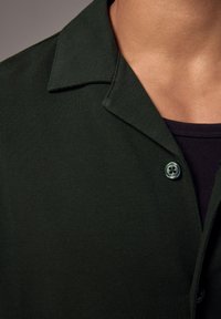 Camicia button-up verde scuro con tessuto texturizzato, colletto corto e un dettaglio con un solo bottone, indossata sopra una maglietta nera.