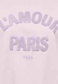 Lettres texturées violet clair formant "L'AMOUR PARIS" avec l'année "1996" brodée en dessous sur un fond de tissu pâle.