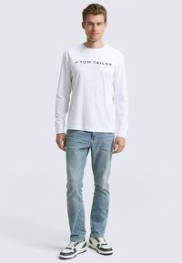 Wit T-shirt met lange mouwen en zwart "TOM TAILOR" logo, gecombineerd met lichtblauwe spijkerbroek en zwart-witte sneakers, staand tegen een neutrale achtergrond.