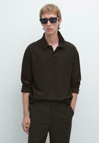 Giovane con capelli castano chiaro che indossa occhiali da sole neri, una maglia a maniche lunghe marrone scuro e pantaloni scuri coordinati, su uno sfondo bianco.