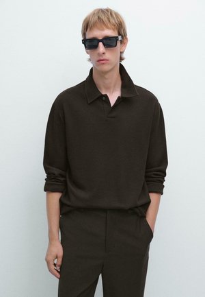 Giovane con capelli castano chiaro che indossa occhiali da sole neri, una maglia a maniche lunghe marrone scuro e pantaloni scuri coordinati, su uno sfondo bianco.