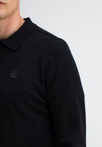 Schwarzes Polohemd aus strukturiertem Stoff. Verfügt über einen klassischen Kragen und ein kleines, geprägtes Logo auf der Brust. Einfaches, schnörkelloses Design.