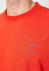 T-shirt en coton rouge avec un col rond, arborant deux logos bleus "SAMSOE" sur la zone gauche de la poitrine, texture lisse et coupe décontractée.