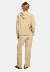 Vrouw staat met haar rug naar de camera, draagt een beige hoodie, bijpassende broek en bruine slippers, haar in een knot.