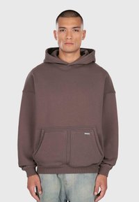 Rövid hajú férfi, aki laza barna kapucnis pulcsit visel elülső zsebekkel és világoskék farmernadrággal, előre néz egy egyszínű háttér előtt.