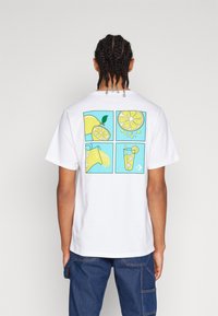 Camiseta blanca de algodón con un gráfico colorido en la parte posterior que presenta cuatro paneles de ilustraciones de limones y limonada sobre un fondo turquesa.