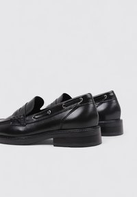 Mocasines de cuero negro con una superficie lisa, que cuentan con costuras decorativas y ojales metálicos, además de un tacón bajo para mayor soporte.